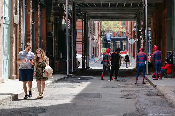A couple walks down a sunny NYC street with coffee while in the background several people dressed as Spidermen hold a meeting.